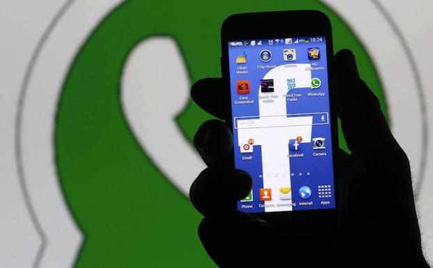 Un usuario sostiene un móvil con el logo de Facebook en la pantalla, ante el icono de Whatsapp./REUTERS